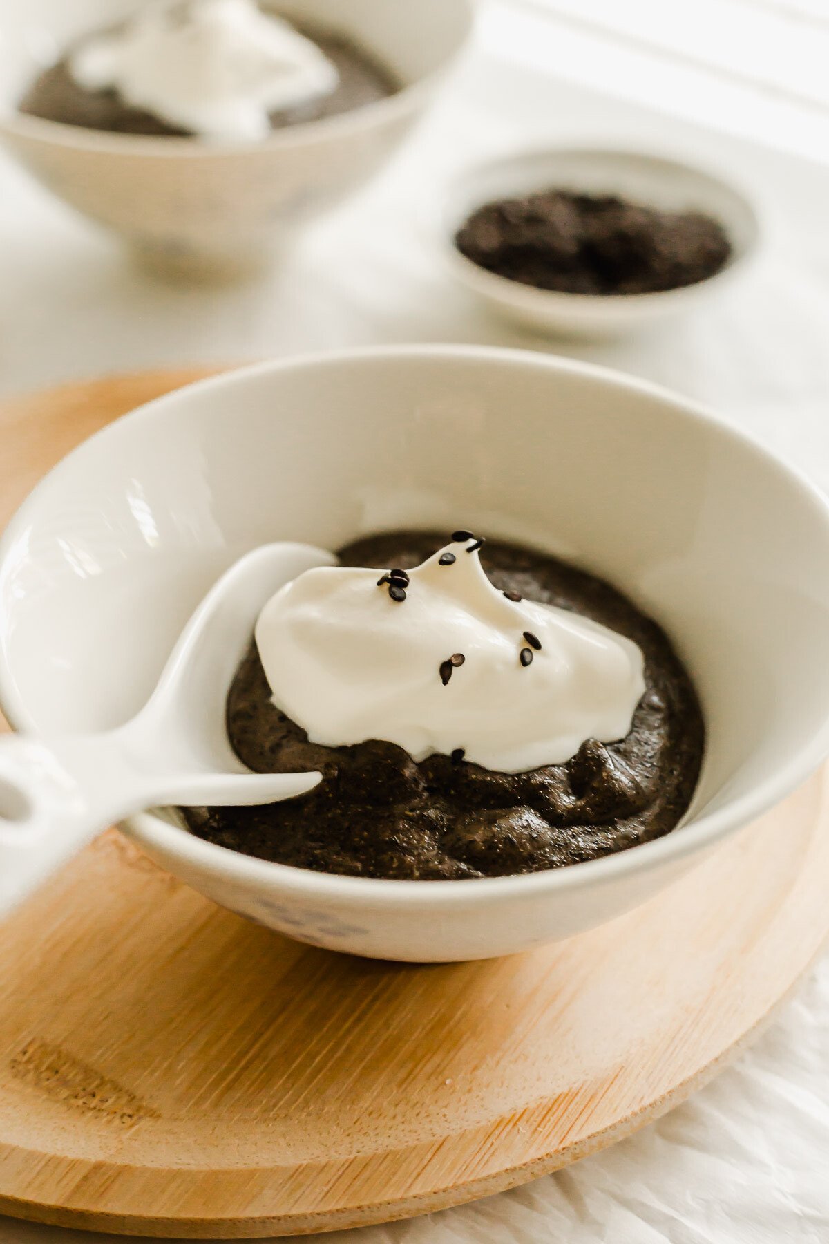 Black sesame paste in a white bowl with spoon, with a dollop of whipped cream and black sesame seeds.